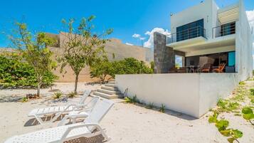 A modern two-story house with a balcony, white lounge chairs, and a sandy landscape.