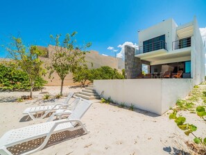 Una casa moderna de dos pisos con un balcón, sillones blancos para relajarse y un paisaje arenoso.