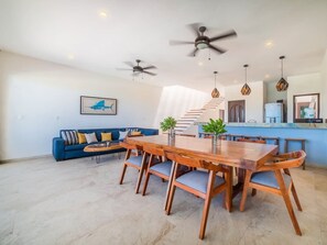 Amplia sala de estar con una mesa de comedor de madera, sofá azul, ventiladores de techo y una escalera que conduce a un nivel superior.