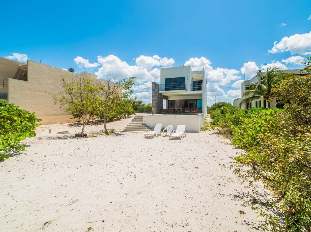 A modern beach house with a two-story structure, a sandy patio with lounge chairs, and a staircase leading to the upper level.