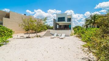 A modern beach house with a two-story structure, a sandy patio with lounge chairs, and a staircase leading to the upper level.