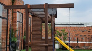 Estructura de madera para juegos con un tobogán y columpios, frente a un muro de ladrillo con plantas en macetas.