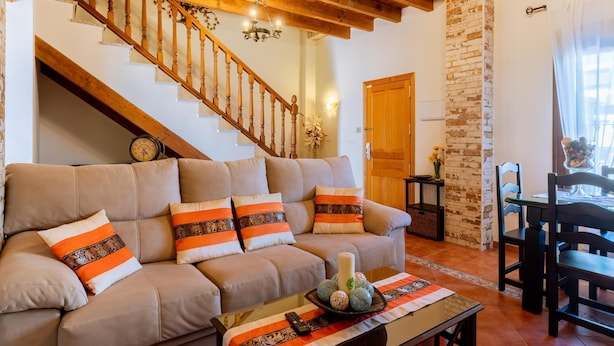 Una sala de estar con una escalera de madera, un sofá con cojines estampados, una mesa de centro con objetos decorativos y un comedor con mesa y sillas.