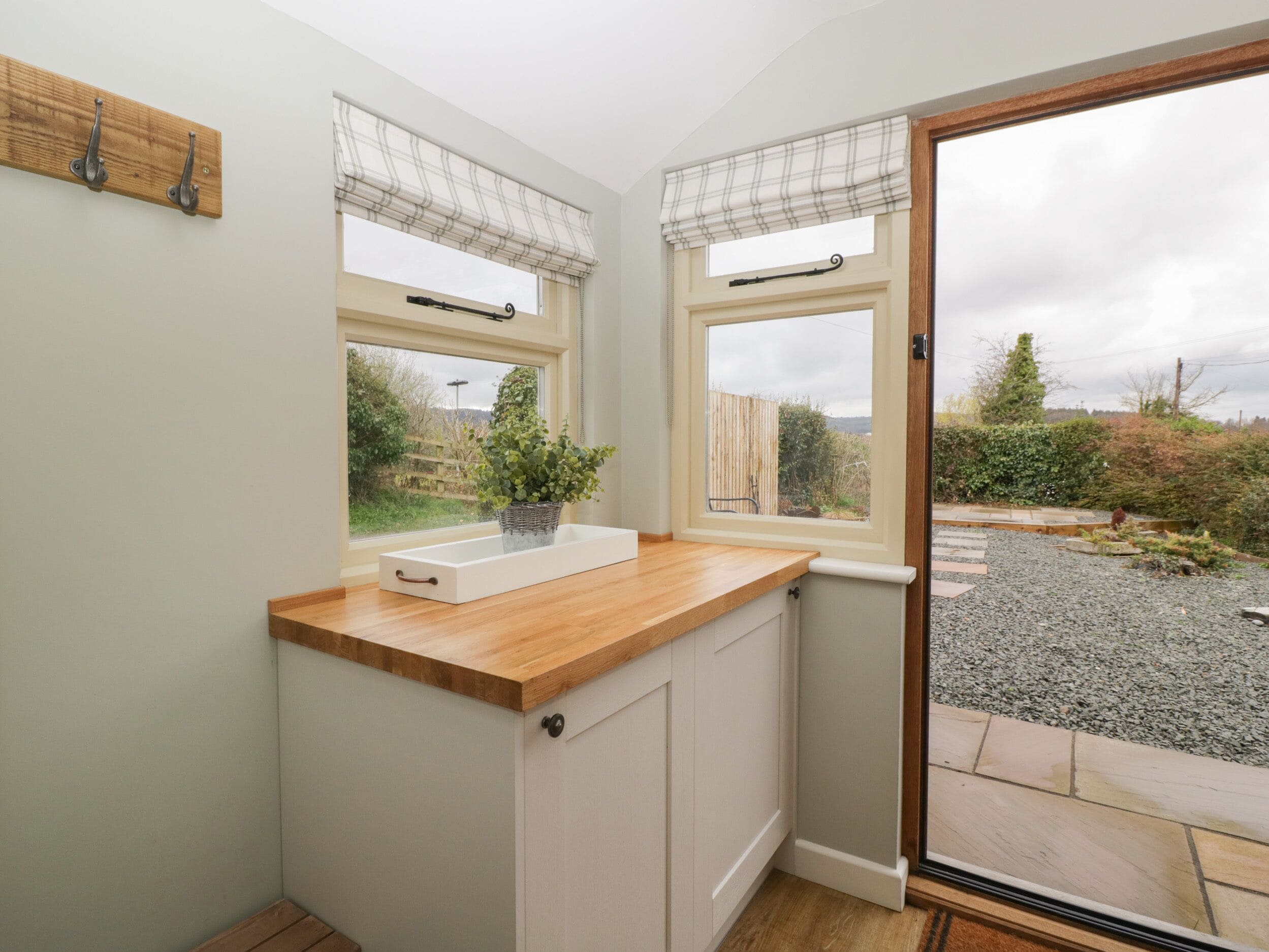 A small kitchen with a wooden countertop, two windows with curtains, a potted plant, and a view of the garden.