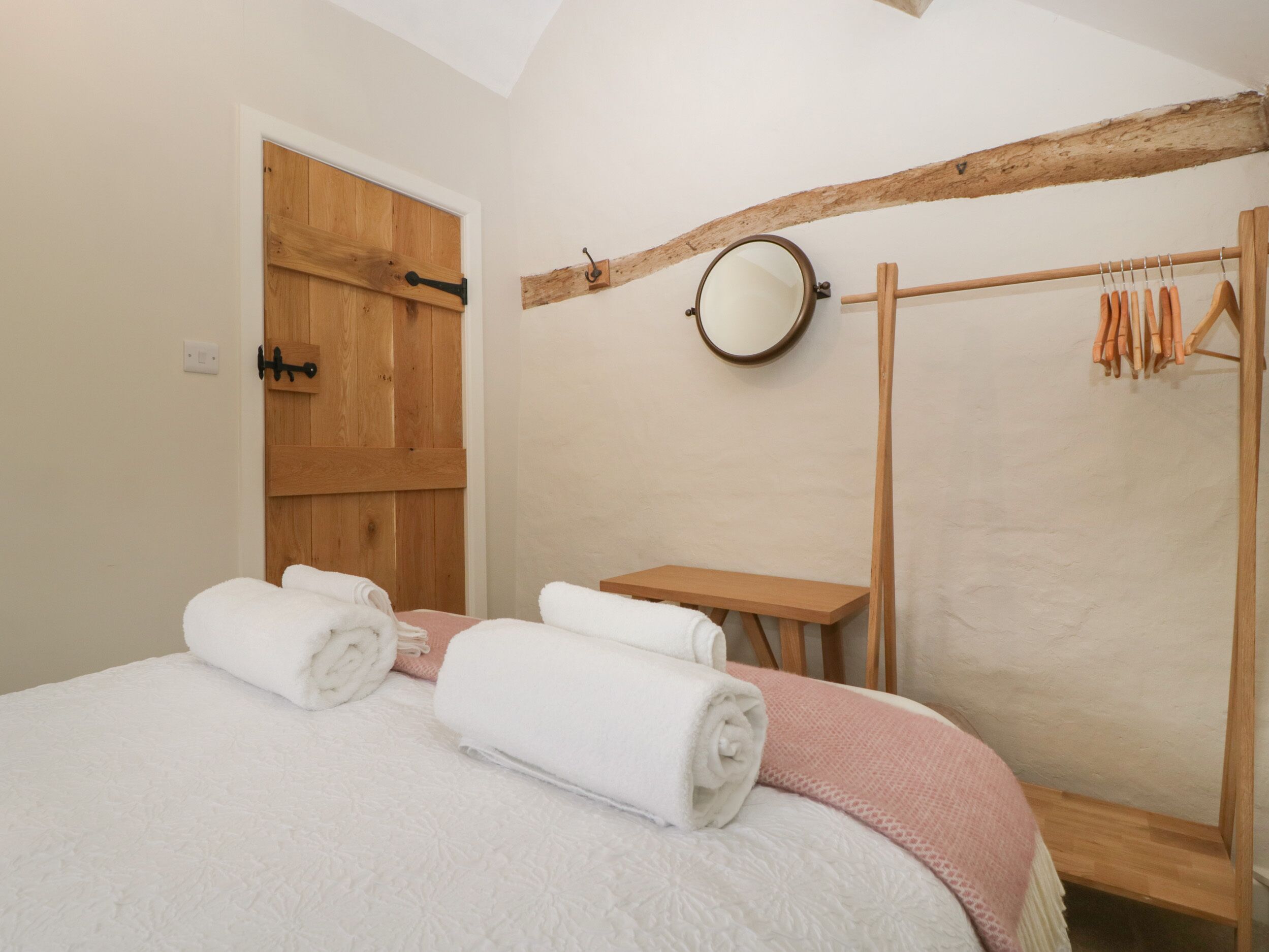 Une chambre avec une porte en bois, un miroir rond, un banc en bois et un portant à vêtements avec des cintres.