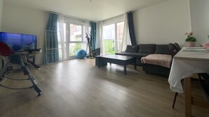 Un salon spacieux avec un canapé, une table basse et un téléviseur. Il y a une grande fenêtre avec des rideaux, un ballon d’exercice bleu et un ventilateur.
