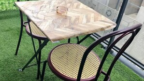 Une table en bois à motifs en chevrons, sur laquelle repose un cendrier en verre, entourée de chaises en osier, le tout installé sur une pelouse artificielle.