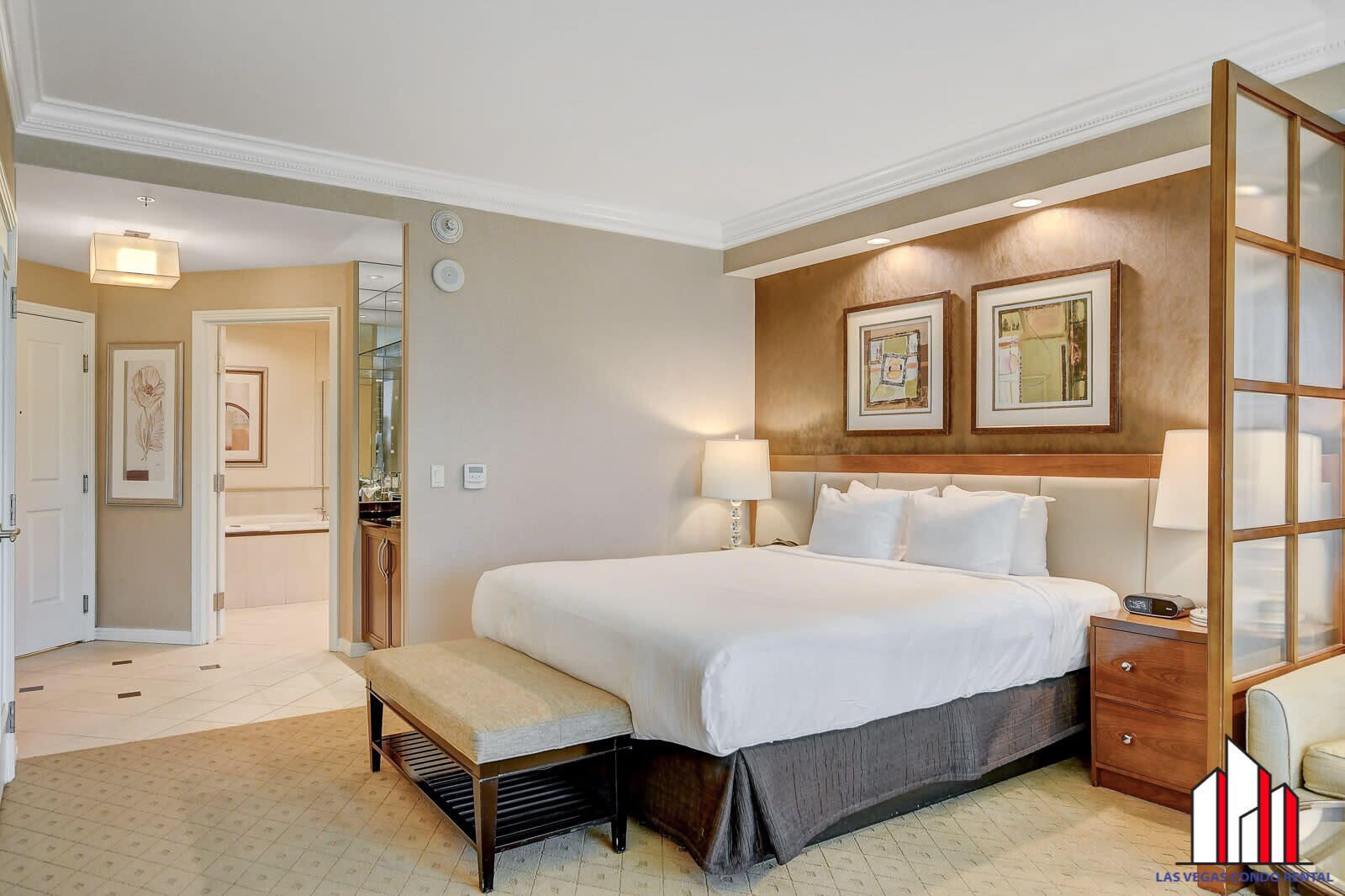 A hotel bedroom with a large bed, bedside tables, a bench, and framed artwork on the wall.
