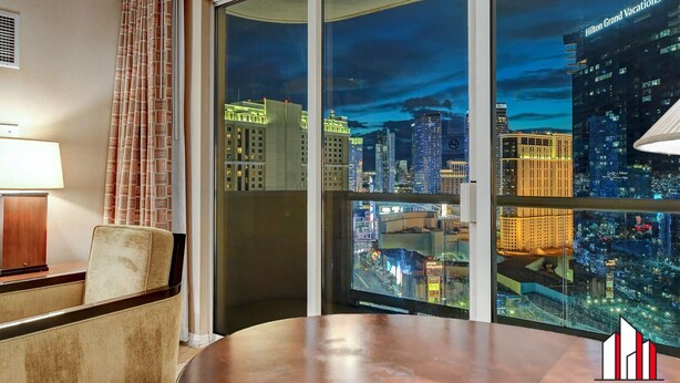 A hotel room with a large window offering a nighttime city view, a round table, a chair, and a lamp.