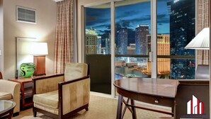 A hotel room with a large window offering a city view, a desk, a chair, a lamp, and a decorative vase.