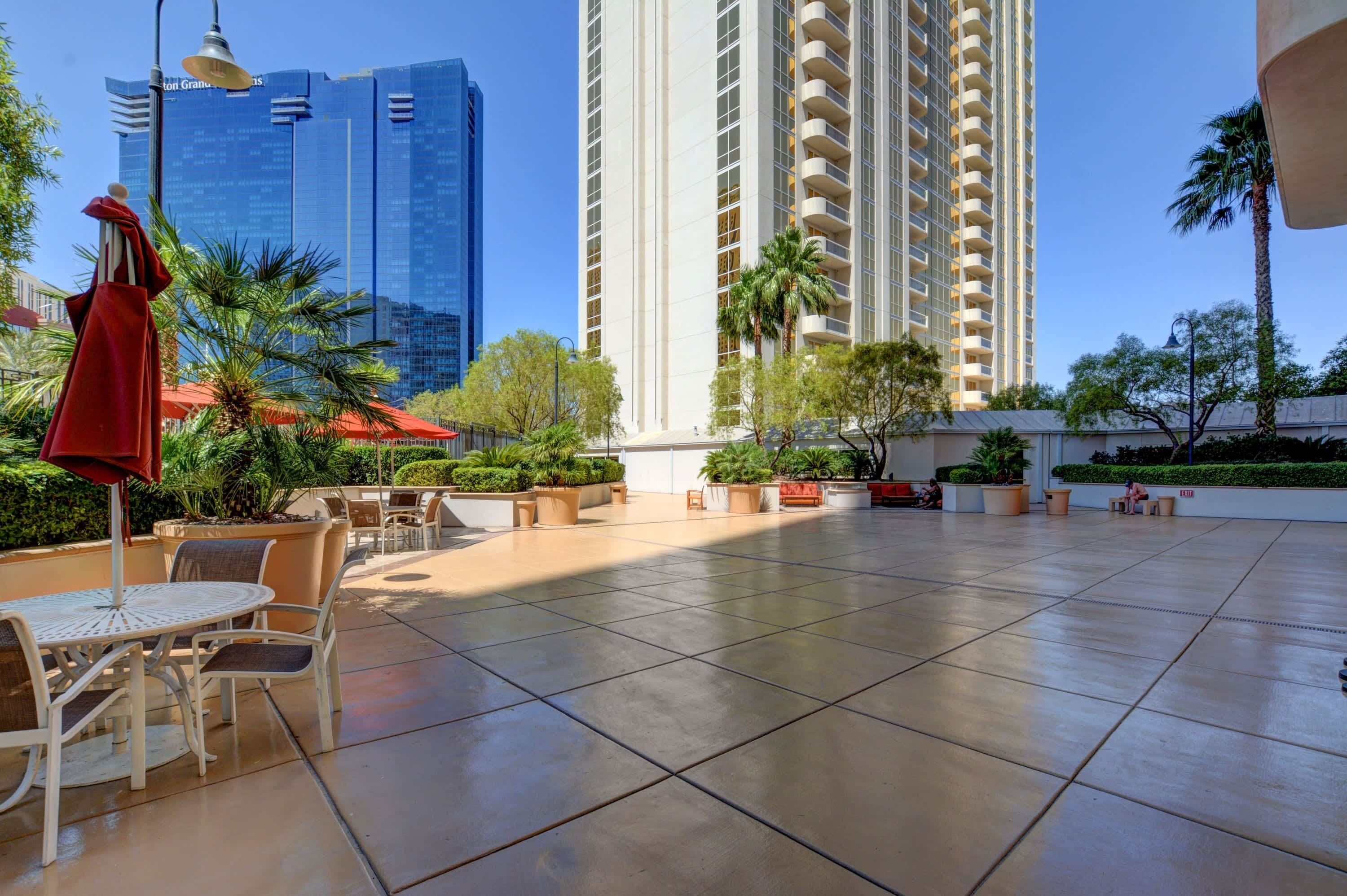 A spacious outdoor patio with a seating area, surrounded by lush greenery and palm trees, with tall buildings in the background.