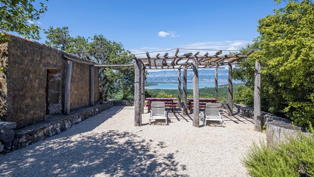 Ein rustikaler Sitzbereich im Freien mit einer Pergola, Steinmauern und Blick auf einen See.