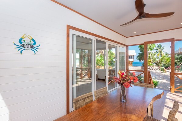 A room with a wooden table, a vase with flowers, and a view of the beach and palm trees through large glass doors. - Casa Coral And Casa Palmera 4 Bedroom Home by RedAwning (Roatan)
