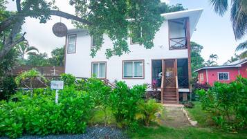A two-story house with a balcony, surrounded by lush greenery and a sign indicating a parking area.