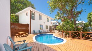 A pool area with a wooden deck, a white building, and a view of the sea. - Casa Coral And Casa Palmera 4 Bedroom Home by RedAwning (Roatan)