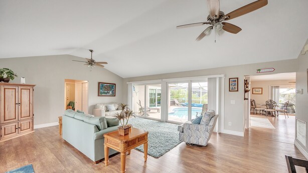 A spacious living room with hardwood floors, a light blue sofa, a patterned armchair, and a wooden coffee table. There are ceiling fans, a large window, and a view of a swimming pool.