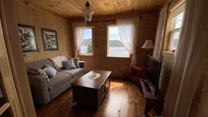 A cozy room with wooden walls and floors, a sofa, a coffee table, a TV, and a window with a view of a lake. - One bedroom two bath beach house over looking the ocean. (Trinity)