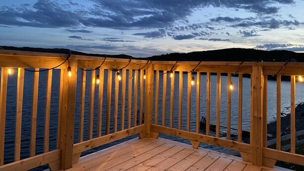 A wooden deck with string lights, overlooking a body of water and a hilly landscape. - One bedroom two bath beach house over looking the ocean. (Trinity)