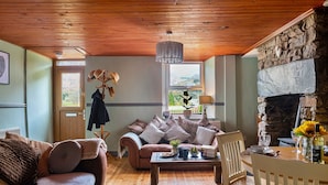 A cozy living room with a wooden ceiling, a stone fireplace, a sofa, a coffee table, and a dining area with chairs and a table set for a meal.