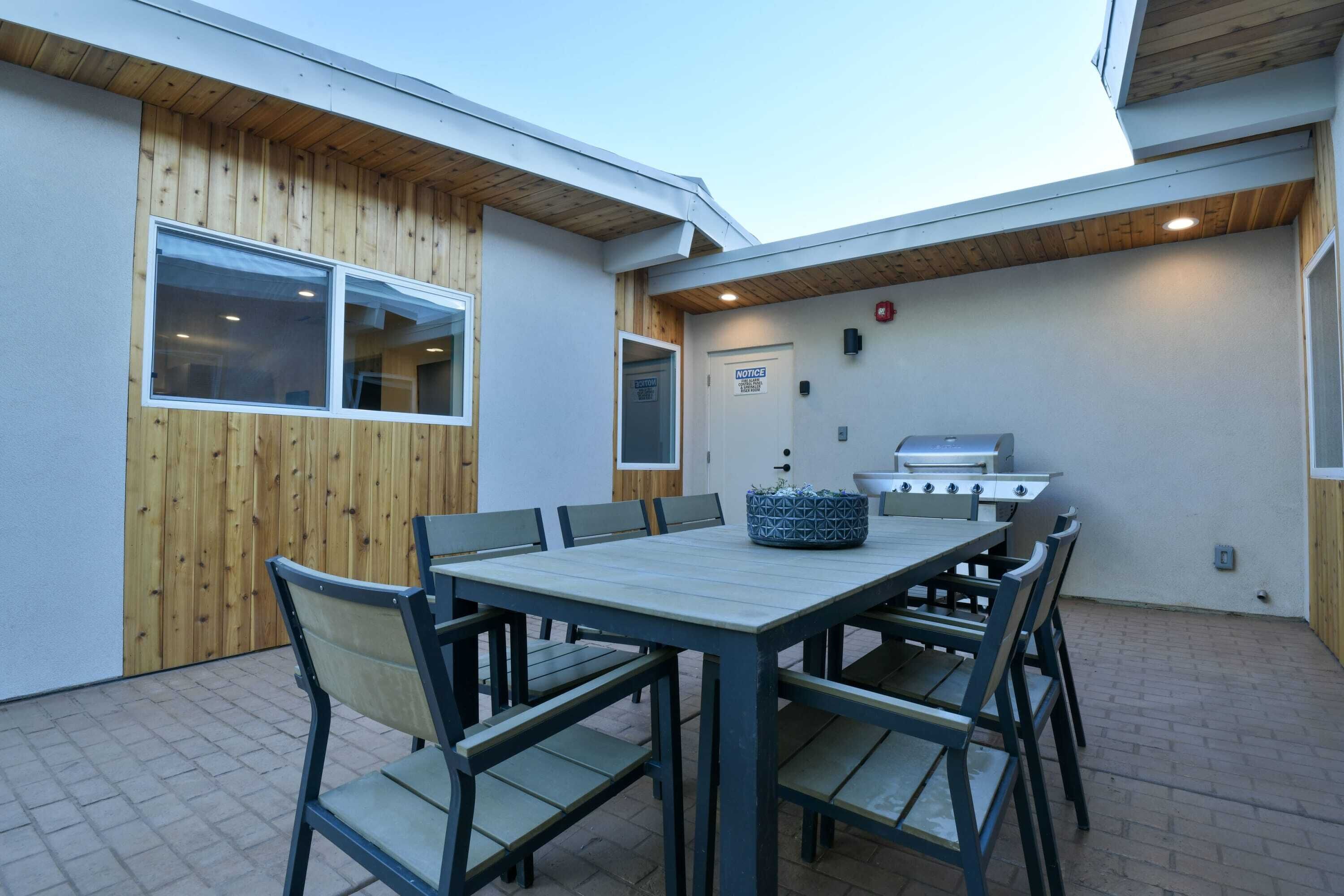 An outdoor patio with a wooden table and chairs, a barbecue grill, and a door with a notice sign.