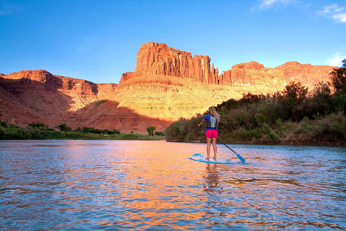 Eine Person steht auf einem Paddleboard auf einem Fluss, im Hintergrund eine gebirgige Landschaft.