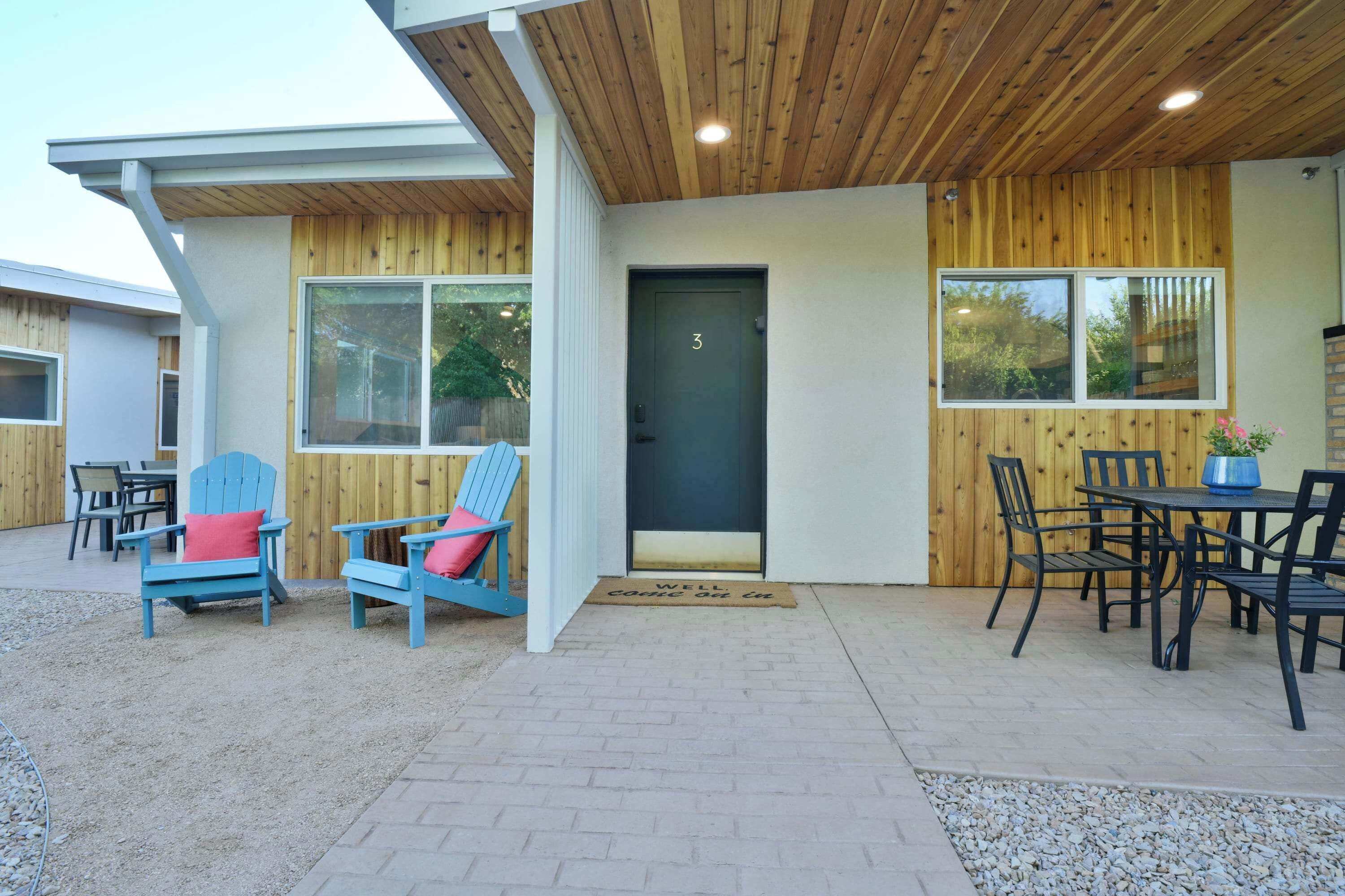 A modern building with a wooden exterior, a black door with the number 3, and outdoor seating with chairs and a table.