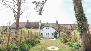 A white house with a thatched roof, surrounded by a wooden fence and lush greenery. - Lime Cottage (Bridport)