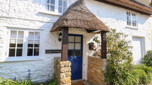 A white building with a blue door and a thatched roof entrance. - Lime Cottage (Bridport)