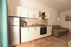 A compact kitchen with white cabinets, a stainless steel refrigerator, a gas stove, and a built-in oven. A brown sofa is placed in the corner. - Apartment Poseidonia - Sole & Pepe - Santa Maria di Castellabate Cilento (Santa Maria di Castellabate)