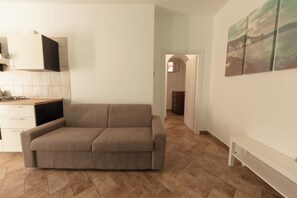 A modern living room with a beige sofa, a white bench, and a kitchen area with a stove and cabinets. - Apartment Poseidonia - Sole & Pepe - Santa Maria di Castellabate Cilento (Santa Maria di Castellabate)