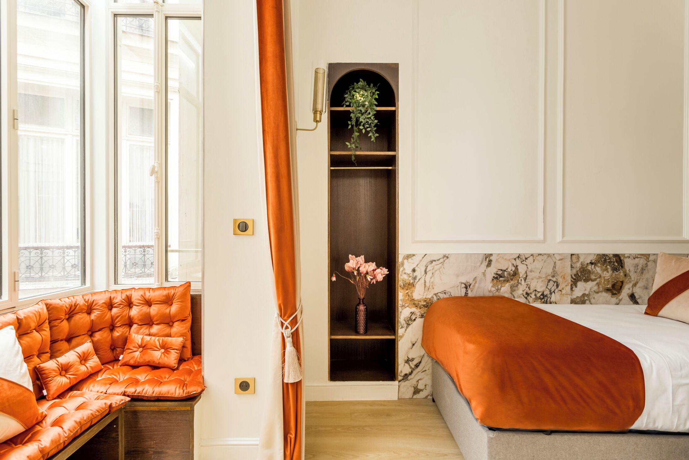 A room with a bed, a cushioned bench, a shelf with a vase, and a window with curtains.