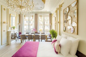 Ein luxuriöses Hotelzimmer mit einem großen Bett, einem Essbereich mit Tisch und Stühlen, einer Kandelaber und großen Fenstern mit Blick auf die Stadt.
