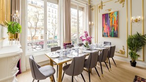 Ein Esszimmer mit großem Tisch, gedeckt für sechs Personen, großen Fenstern mit Vorhängen, einer Kristallleuchtreiter und einem farbenfrohen, abstrakten Gemälde an der Wand.