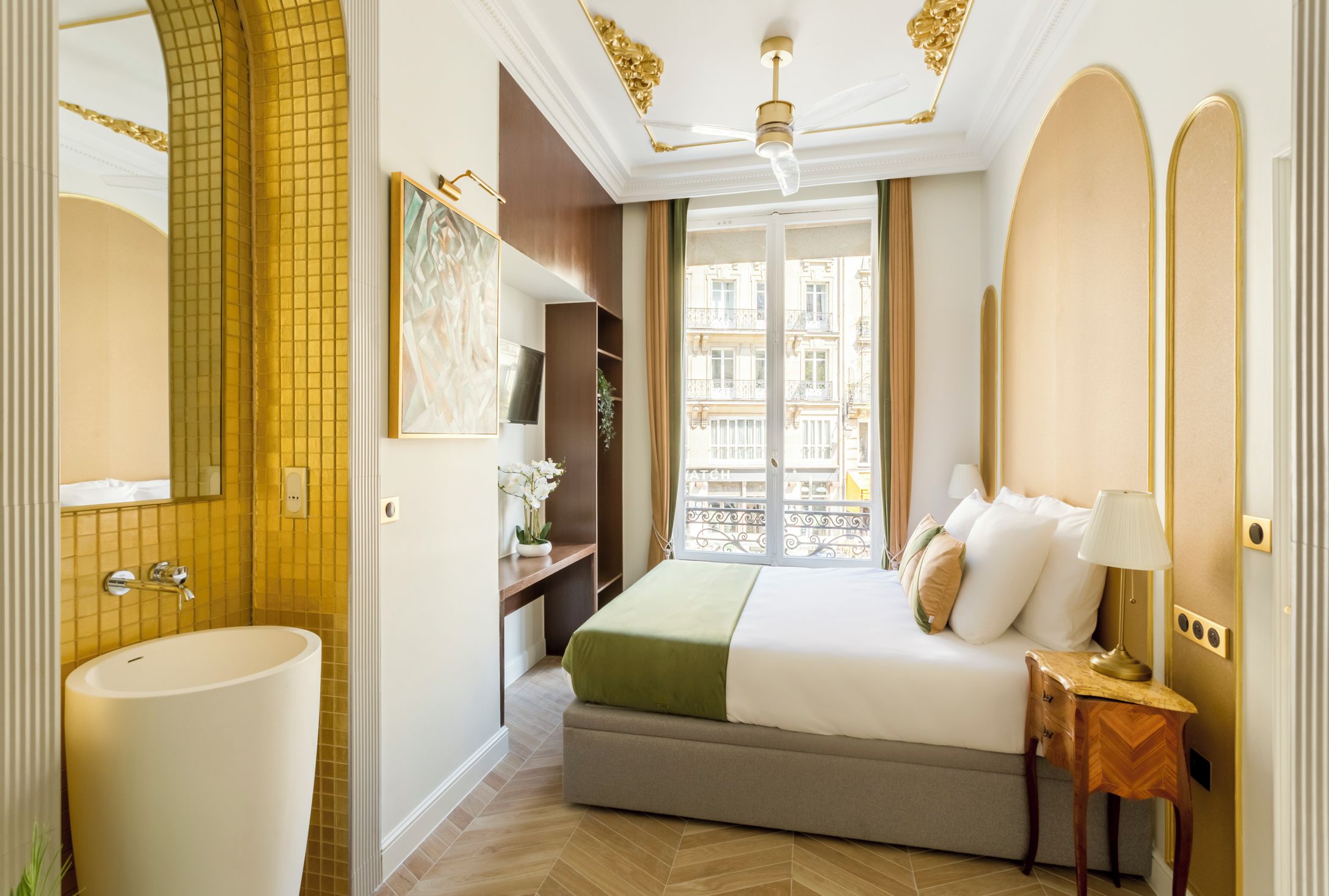 A hotel room with a large bed, a bedside table with a lamp, a large window with a view of buildings, a bathroom with a sink and mirror, and a ceiling fan.