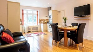 A modern living room with a black leather sofa, a dining table with chairs, a kitchen area with appliances, and a mounted TV. - Sapphire - Spacious apartment near the Sea (Caister-on-Sea)