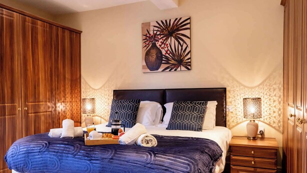 A bedroom with a large bed, wooden wardrobe, bedside tables with lamps, and a wall art piece. - Sapphire - Spacious apartment near the Sea (Caister-on-Sea)
