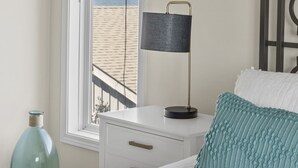 A bedroom with a bed, a nightstand, a lamp, a window with a view of the beach, and a decorative glass bottle. - The Hamptons, Oceanfront Duplex in Atlantic Beach, NC! (Atlantic Beach)