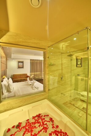 Ein Hotelbad mit einer gläsernen Duschkabine, einer Badewanne mit Rosenblüten, einem Bett mit weißen Bettwäsche und einem gerahmten Bild an der Wand.