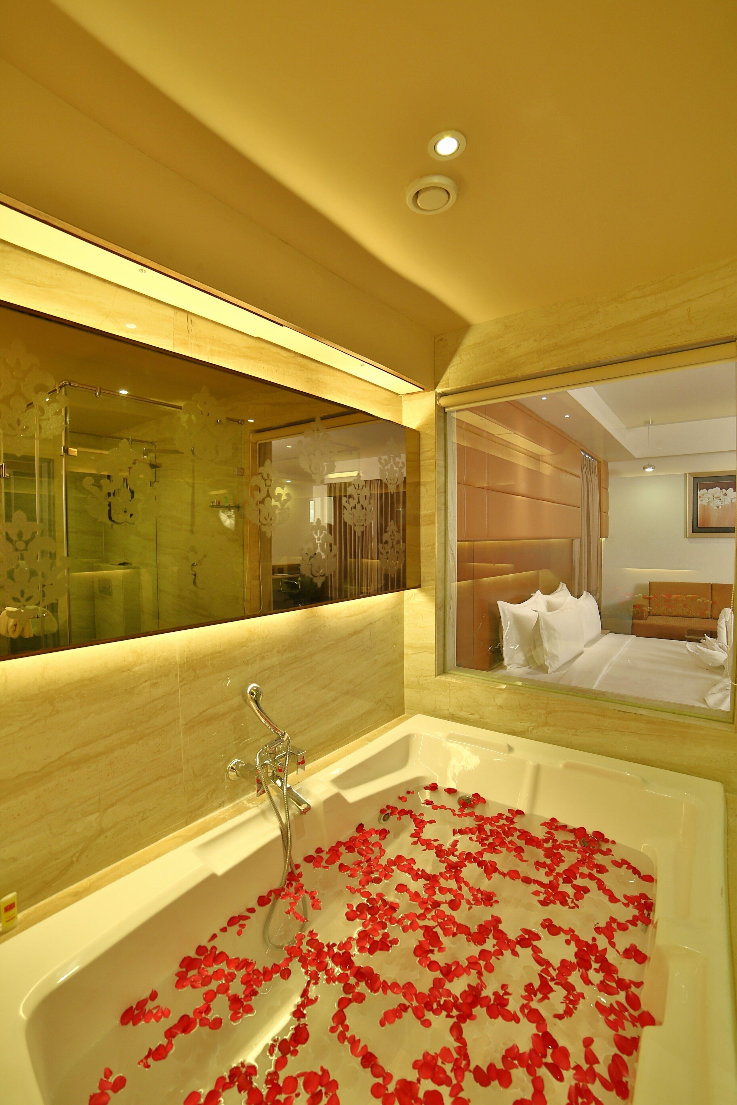 A hotel bathroom with a large bathtub filled with rose petals, a mirror, and a bed visible in the reflection.