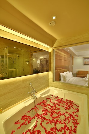 A hotel bathroom with a large bathtub filled with rose petals, a mirror, and a bed visible in the reflection. - hotel G Express (Ahmedabad)