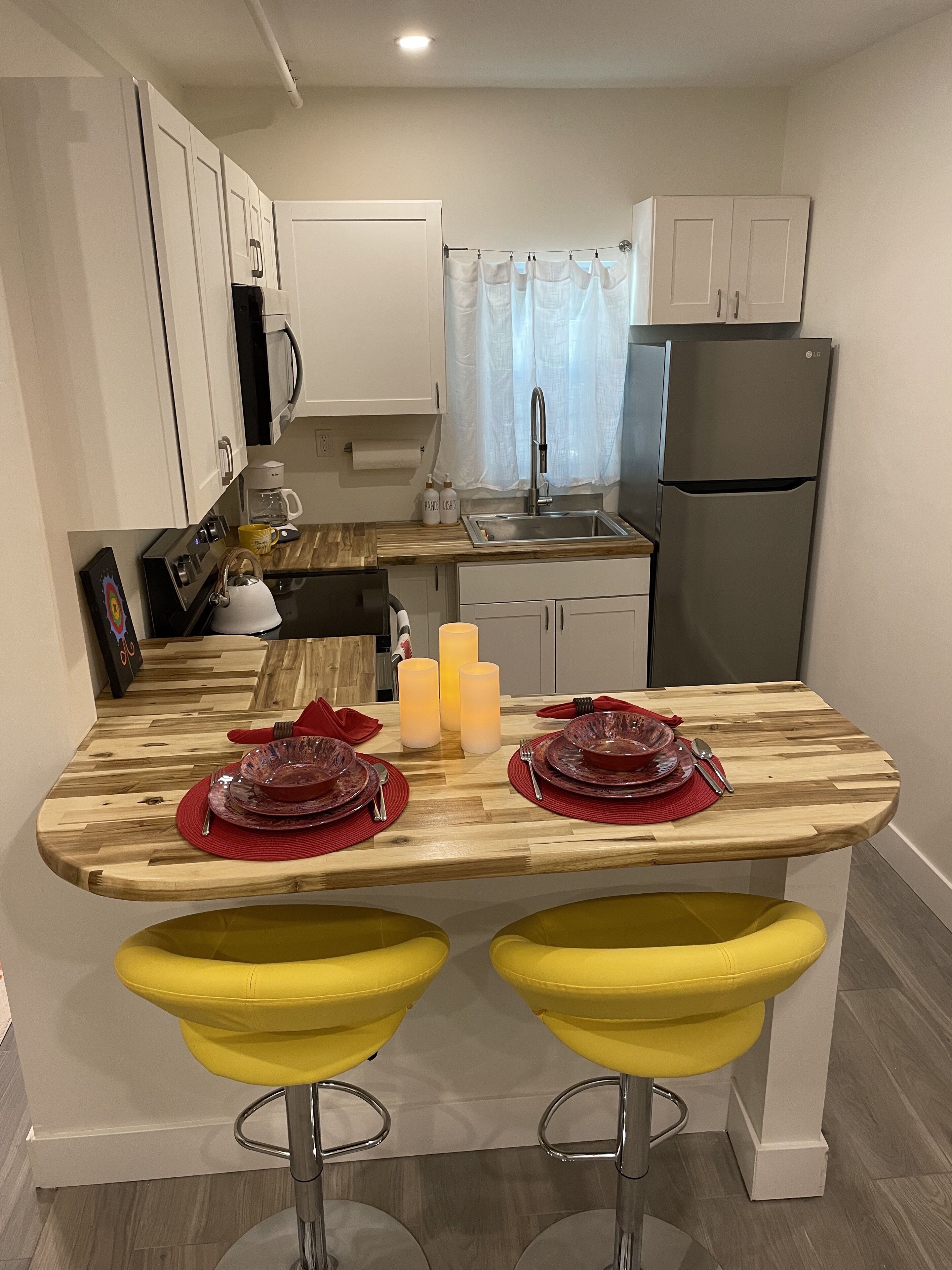 A modern kitchen with white cabinets, a stainless steel sink, and an LG refrigerator. There are two yellow bar stools and a wooden dining table set with red plates and candles.