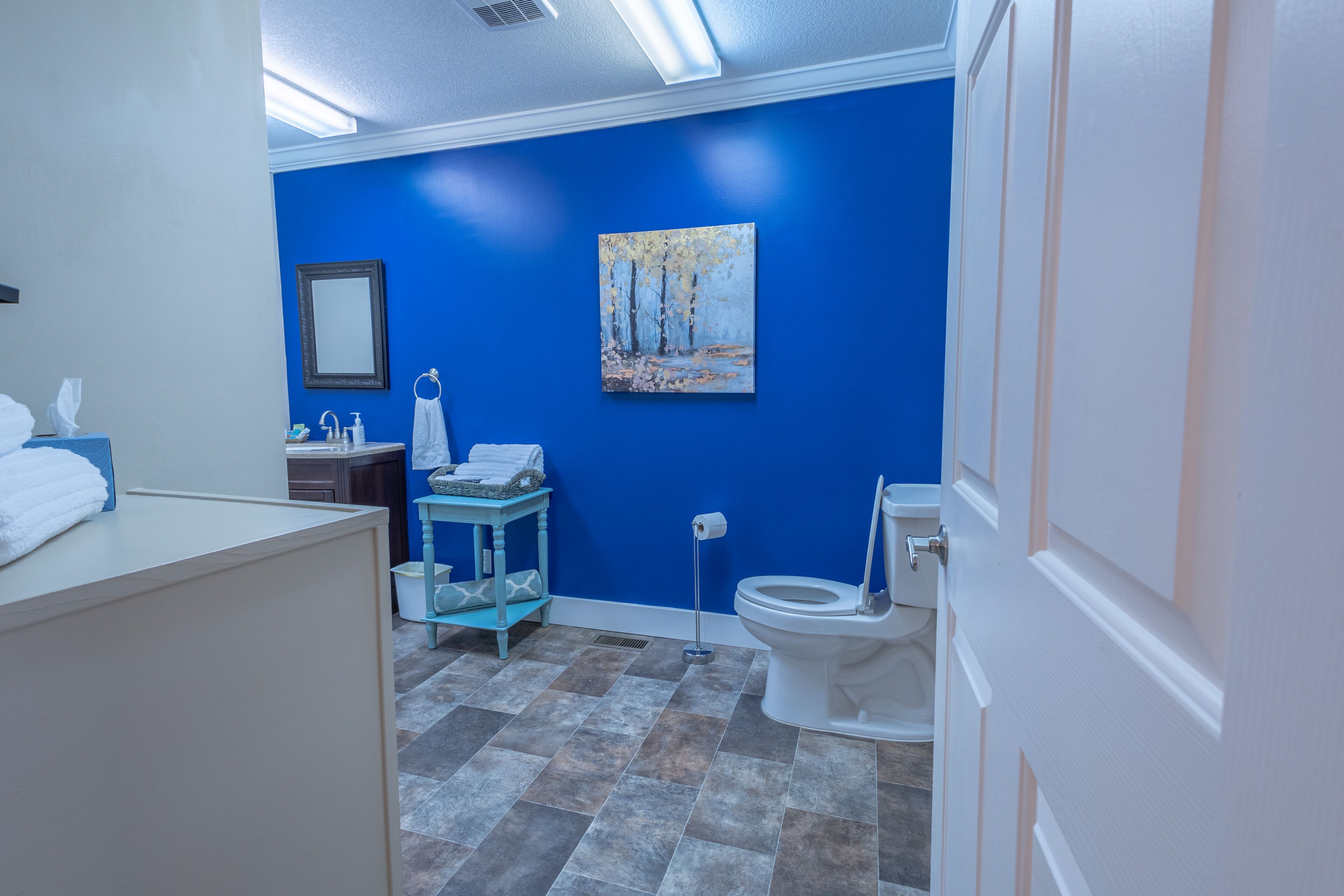 Ein Badezimmer mit blauer Wand, einem Baumgemälde, einer Toilette und einem Waschbecken mit Handtuchhalter.
