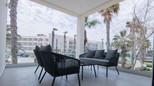 Un balcon avec deux fauteuils en osier noir et un canapé gris, donnant sur un parking et des palmiers.