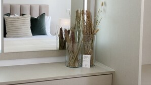 Une chambre à coucher bien rangée, avec un lit, des oreillers, une table de chevet ornée d’un vase contenant des plantes séchées et un miroir qui reflète la pièce.