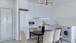 Une cuisine moderne avec des meubles blancs, un réfrigérateur Bosch, un four à micro-ondes et un lave-linge. On y trouve une table à manger avec six chaises et un vase contenant des fleurs séchées.