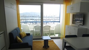 Un espacio habitable compacto con sofá, mesa de comedor y zona de cocina. Un amplio ventanal ofrece vistas a un puerto deportivo con barcos.