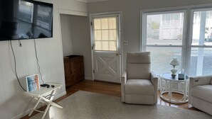 A living room with a flat-screen TV mounted on the wall, a wooden cabinet, a white armchair, a round side table with a lamp, and a sofa. - Waterfront Rancher in South Bethany (Bethany Beach)