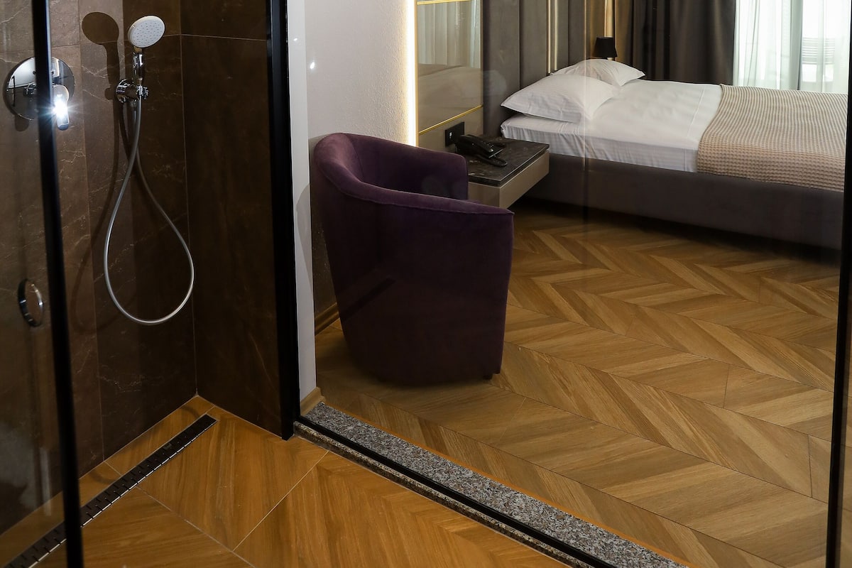 Ett modernt hotellrum med en duschkabin i glas, ett trägolv med fiskbensmönster, en säng med vita sängkläder och en stor spegel som reflekterar rummet.