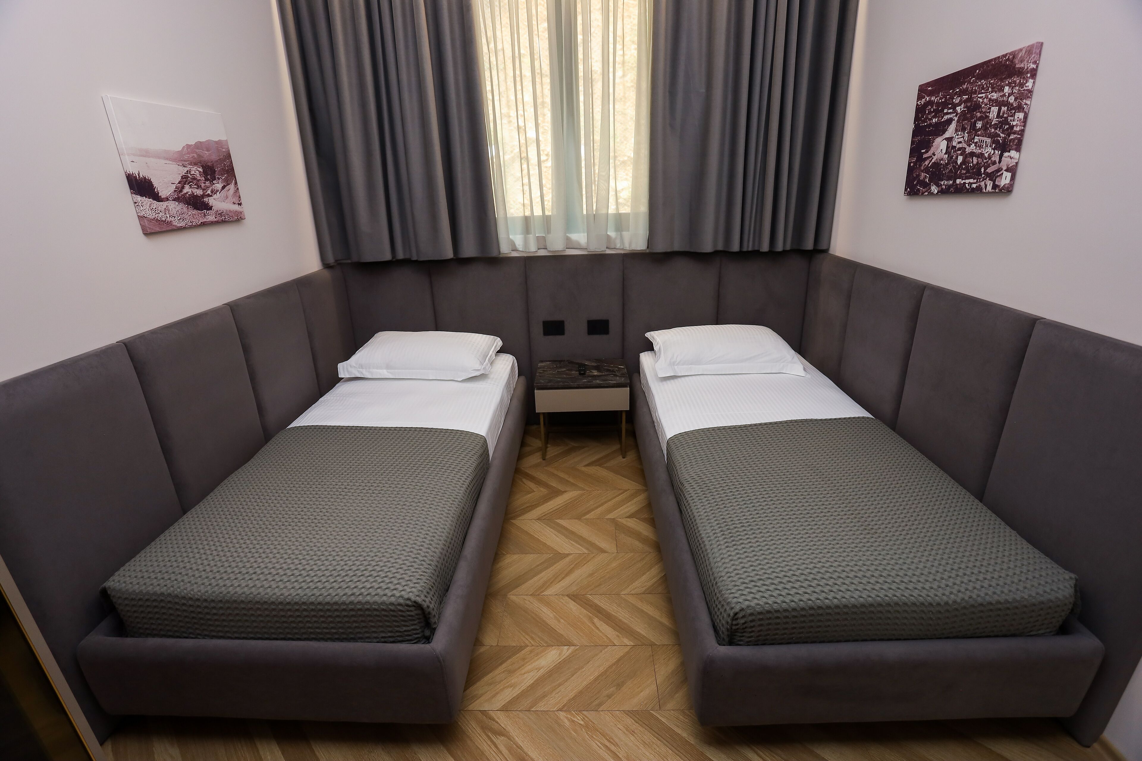 Una habitación con dos camas individuales, un ventanal con cortinas y dos cuadros colgados en la pared.