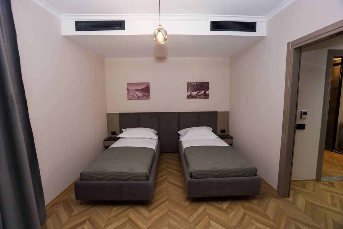 Ett rum med två enkelsängar, ett trägolv med fiskbensmönster, en hängande taklampa och två väggmonterade ventilationsdon.